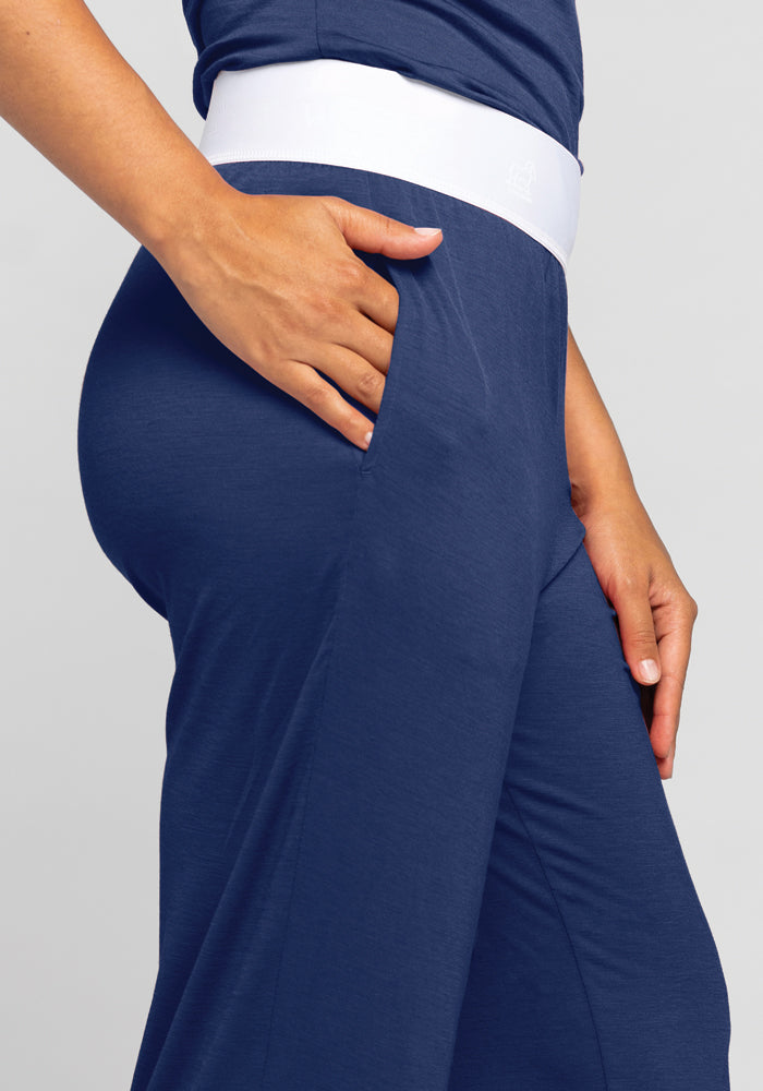 A person wearing the Maya Lounge Pants - Starry Night by Woolx, showcasing a comfortable white waistband, is depicted from the side. They have their right hand tucked into the pant pocket, set against a plain and light-colored background.