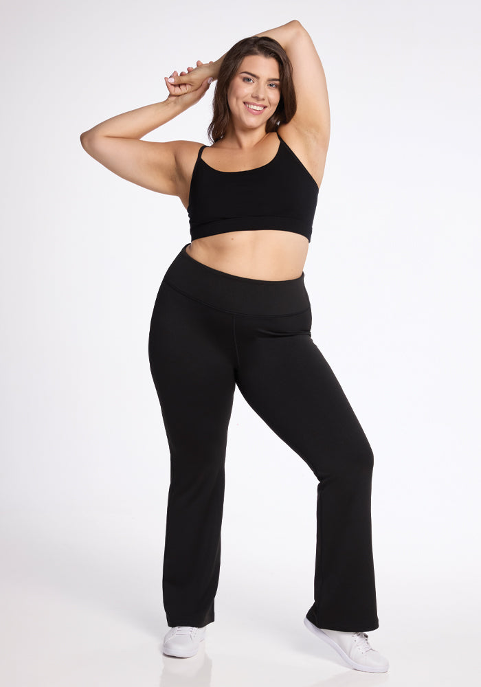 A person is smiling and posing confidently in a black sports bra and Woolx's Marissa Pants - Black. They are standing with one hand on their head and the other on their hip. White sneakers complement the outfit against a plain white background.