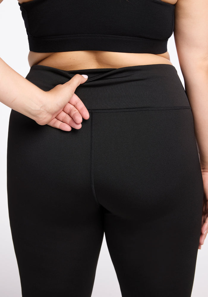 Wearing the Woolx Marissa Pants in black, a person adjusts the waistband at the back. They are facing away from the camera, using one hand to gently pull the waistband. The background is plain with a light color.