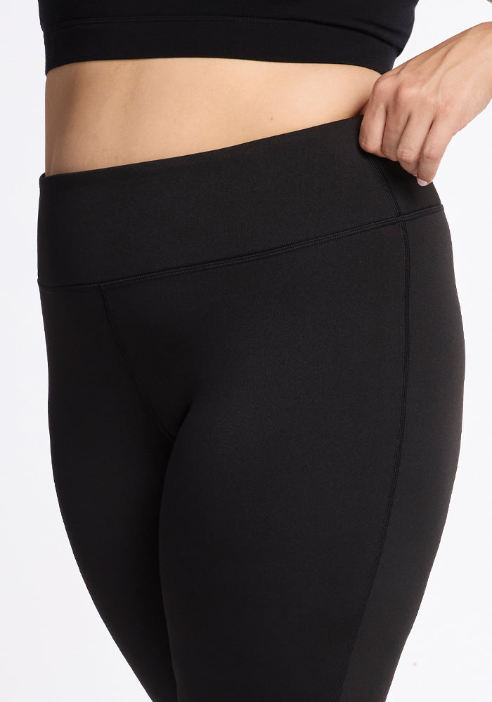 The image features an individual wearing the Marissa Pants in black from Woolx, shown from the waist down. They are adjusting the waistband with their right hand against a plain white background.