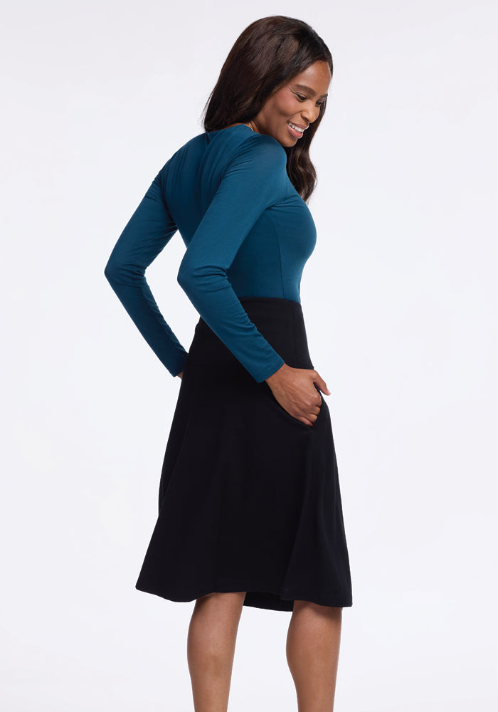 A woman in a long-sleeved teal top and a Woolx Marilyn Flowy Midi Skirt - Black faces sideways, smiling with her hands in the skirt pockets. The flowy design of the skirt adds an elegant touch to her look.