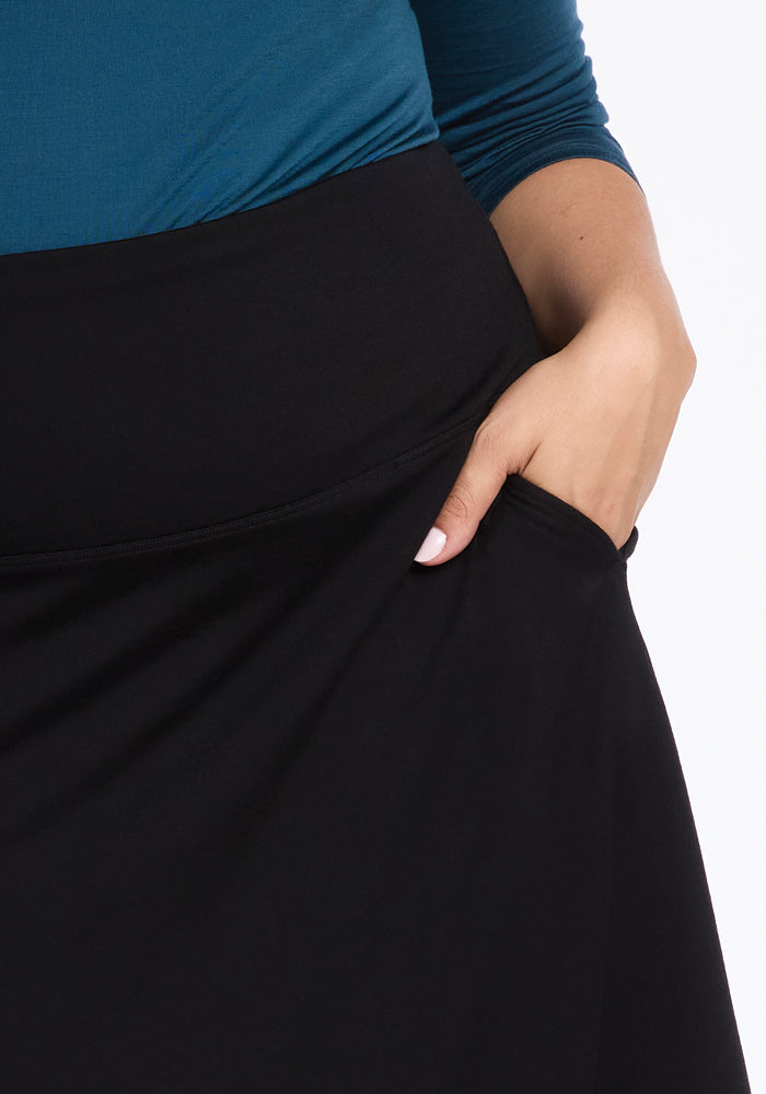 A person wearing a blue top and the stylish Marilyn Flowy Midi Skirt in black by Woolx, with its graceful design, casually tucks a hand into the skirt's pocket. The image beautifully showcases the exquisite detail of this Merino wool creation.