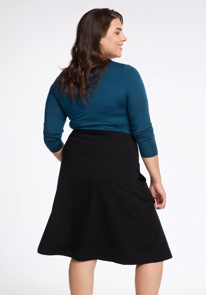 A woman with long brown hair is standing and smiling, wearing a blue long-sleeve top and a Black Marilyn Flowy Midi Skirt from Woolx, known for its elegant flowy design. She is facing away from the camera against a plain, light background.