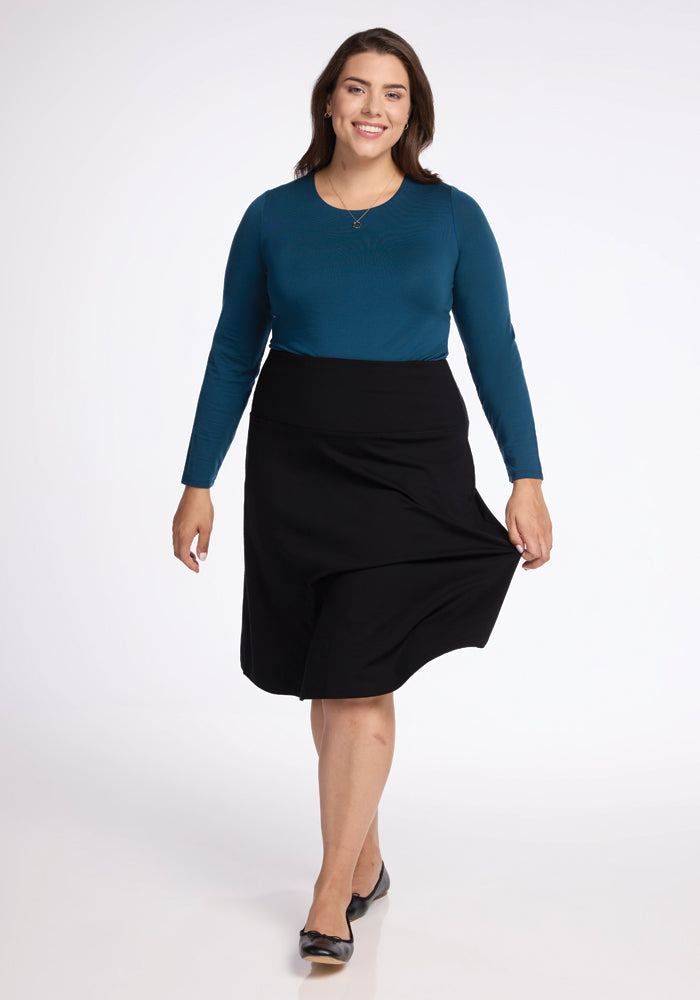 A woman with long brown hair is smiling and walking towards the camera, gracefully donning a teal long-sleeve top and the elegant Woolx Marilyn Flowy Midi Skirt in black, known for its flowy design. Her black flats complete the look against a plain white background.