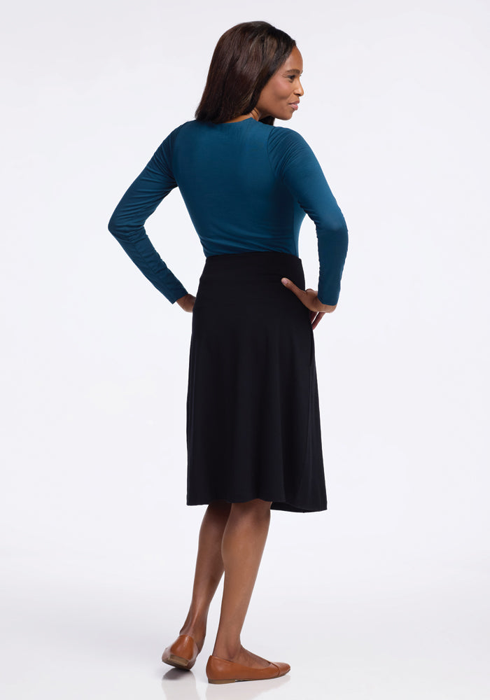 A woman stands with her back to the camera, hands on hips, wearing a teal long-sleeve top paired with a Marilyn Flowy Midi Skirt in Black by Woolx. Her ensemble is finished with brown flats against a plain white background.