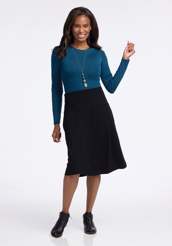 A person stands against a plain white background, smiling and wearing a flowy, long-sleeved teal top and the Marilyn Flowy Midi Skirt in Black from Woolx, paired with black ankle boots. They have long, wavy hair and hold one hand up slightly.