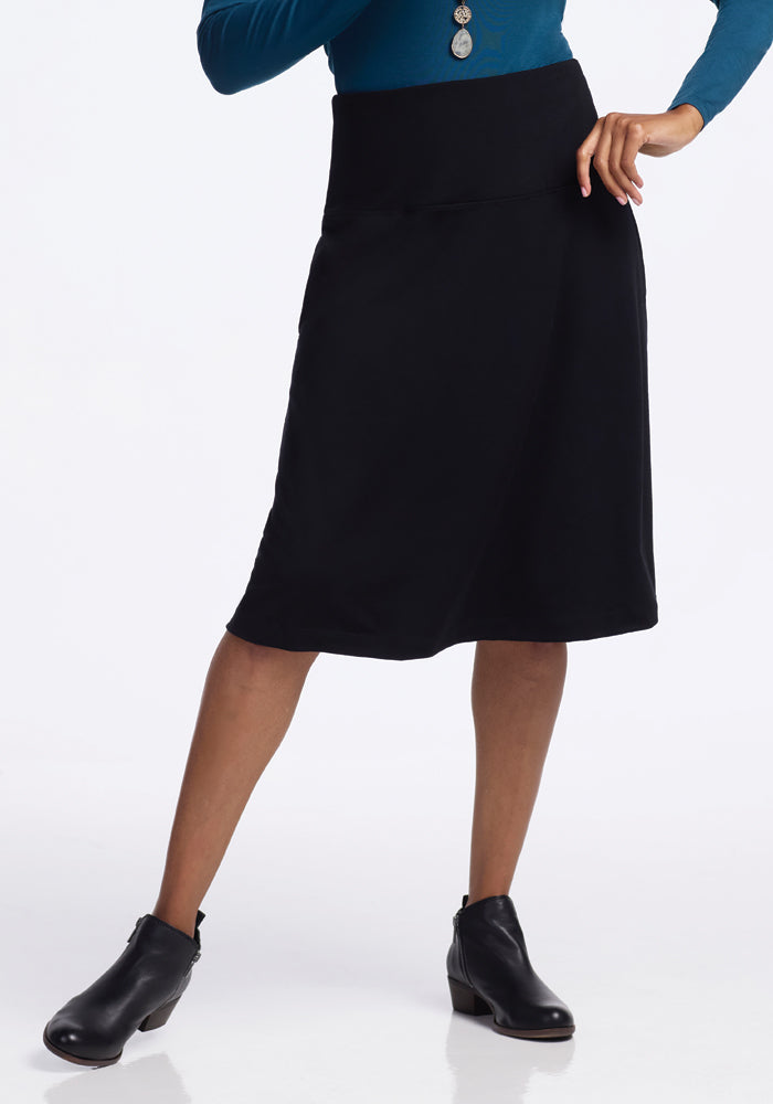 A person wearing a blue long-sleeve top and the Woolx Marilyn Flowy Midi Skirt - Black stands confidently in black ankle boots, one hand on their hip.