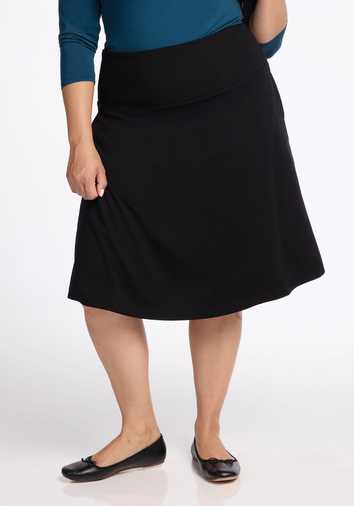 A person wearing a blue top and the Marilyn Flowy Midi Skirt in Black by Woolx, featuring its flowy design, stands on a white background. They are also sporting black ballet flats, with the image cropped from the shoulders down.