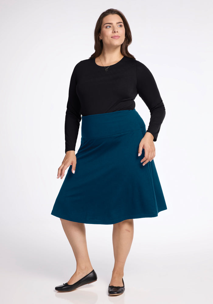 A person stands in a relaxed pose against a plain background, wearing a black long-sleeve top and the Marilyn Flowy Midi Skirt in Real Teal by Woolx, paired with black flats. Their hair is down and styled naturally.