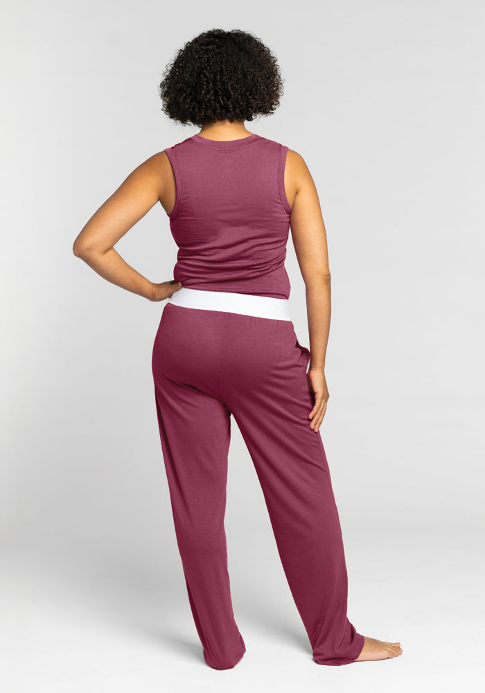 A person with curly hair is depicted from behind wearing a sleeveless maroon top and the Woolx Maya Lounge Pants in Wild Ginger, which feature a comfortable waistband. The ensemble stands out against the plain gray background.