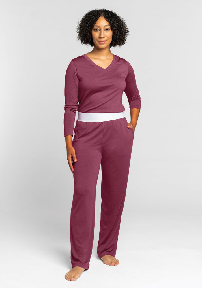 A person with curly hair is dressed in a maroon long-sleeve top and the matching Maya Lounge Pants in Wild Ginger by Woolx, which feature a comfortable waistband. They are standing against a plain gray background, barefoot with one hand in their pocket.