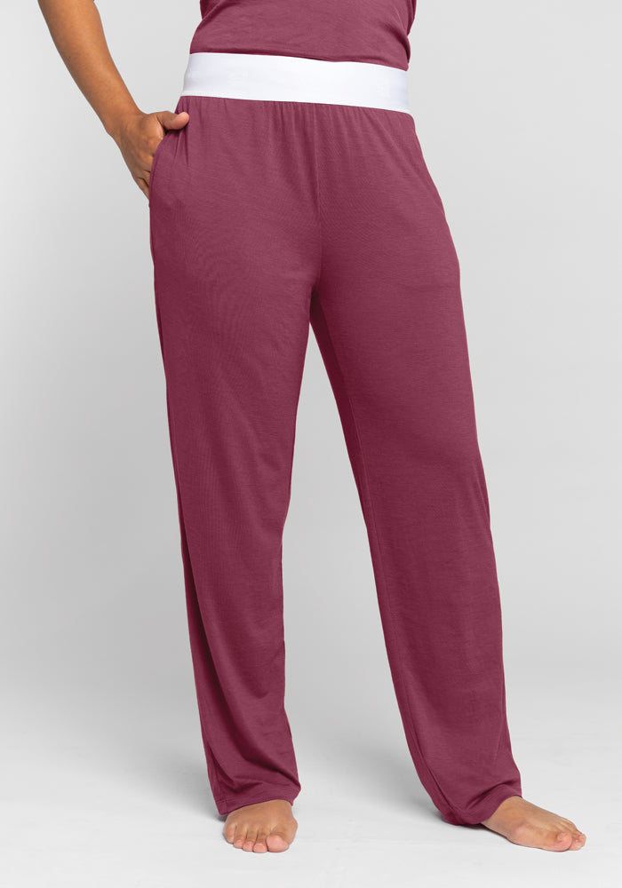 The person is dressed in Woolx Maya Lounge Pants in Wild Ginger, made from lightweight fabric and highlighted by a white waistband. These relaxed-fit pants flow comfortably as the individual stands barefoot with one hand in the pocket against a simple background. 