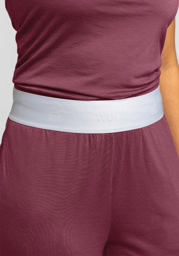 A person is wearing maroon athletic attire, featuring a sleeveless top and the Maya Lounge Pants in Wild Ginger by Woolx, complete with a comfortable white waistband crafted from Merino Wool. The background is plain white.