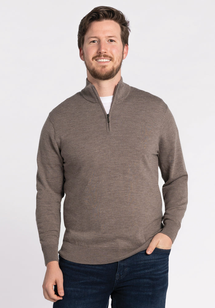 A man wearing the Enzo Quarter Zip in Sable from Woolx, paired with dark jeans, stands against a plain white background. He is smiling with one hand in his pocket. 