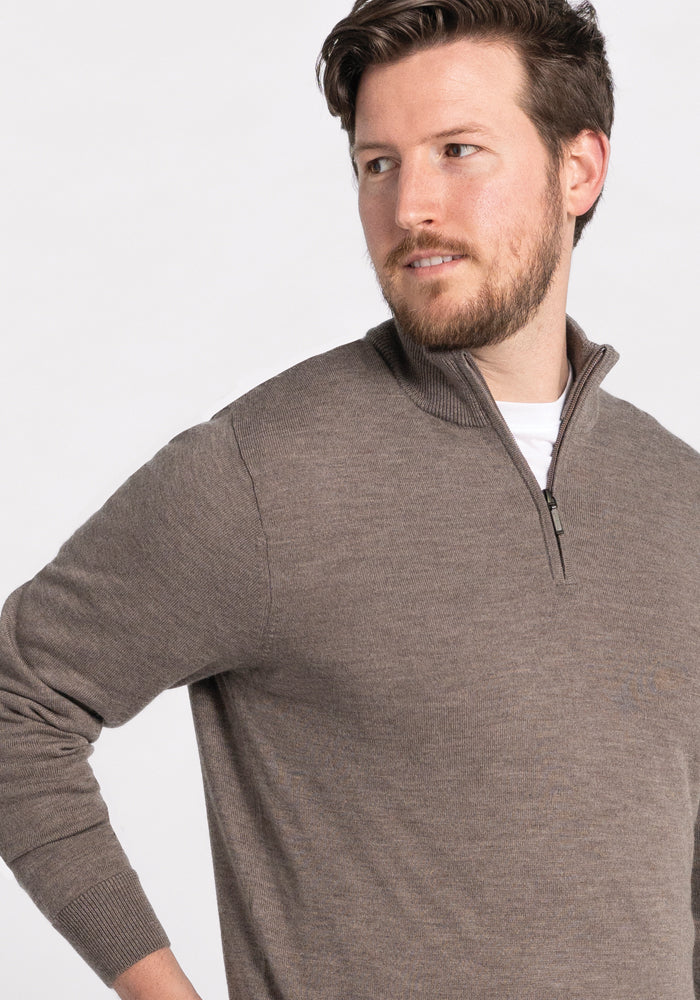 A man with short hair and a beard is wearing the Enzo Quarter Zip in Sable from Woolx over a white shirt. He is looking off to the side against a plain background.