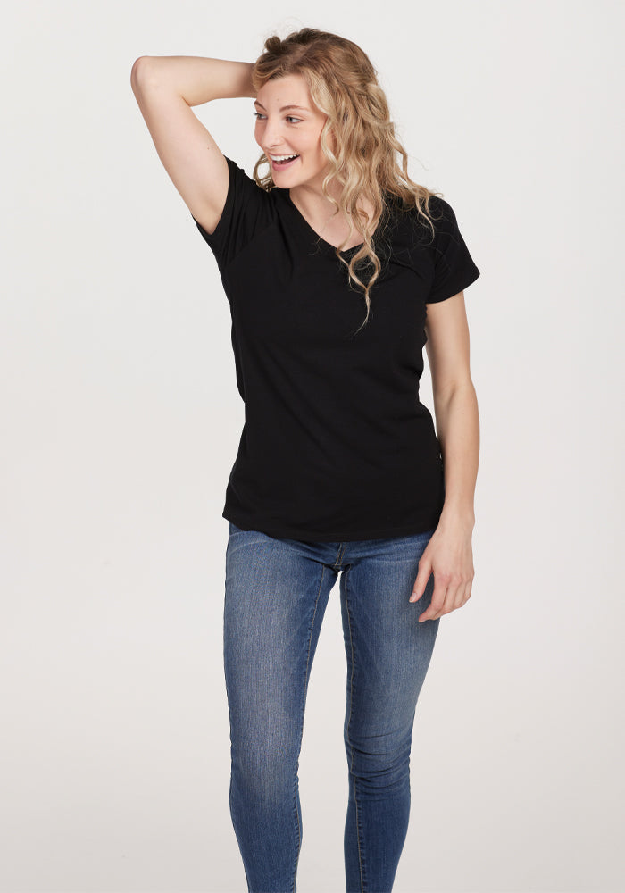 A person with long hair smiles, standing with one hand on their head, wearing the Mia V Neck in black by Woolx and blue jeans against a plain background.