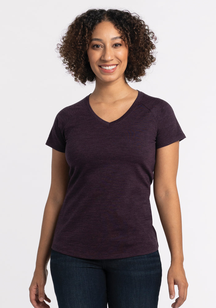 A person with curly hair smiles while standing against a plain background. They are wearing the Woolx Mia V Neck in Deep Plum, a stylish merino wool shirt that's odor-free, paired with dark jeans. 