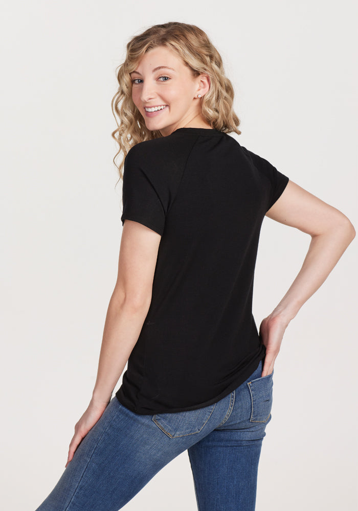 A person with long, wavy blonde hair is smiling and looking over their shoulder. They are wearing the Woolx Mia V Neck - Black, a superior quality women's merino wool tee, along with blue jeans, standing against a plain white background.