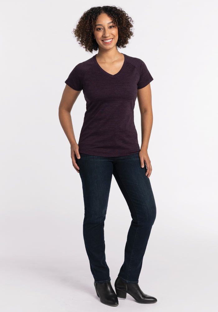 A person with curly hair is smiling and standing against a plain white background. They are wearing a dark purple Mia V Neck - Deep Plum from Woolx, dark jeans, and black boots. Their arms are relaxed at their sides.