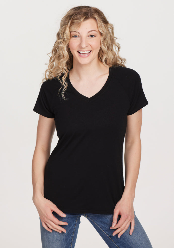 A person with curly blonde hair is smiling while wearing the women's Mia V Neck in black by Woolx, crafted from premium merino wool, paired with blue jeans. They are standing against a plain white background. 