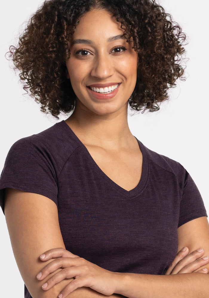 A person with curly hair smiles warmly, standing with arms crossed. They're wearing the deep plum Mia V Neck by Woolx. The plain white background highlights their relaxed and friendly demeanor, showcasing the comfort and style that makes this tee perfect for every adventure.