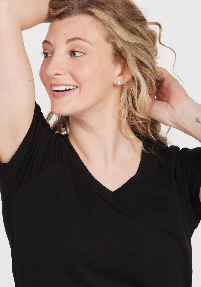 A person with wavy blonde hair smiles while looking to the side, wearing the high-quality Mia V Neck - Black shirt from Woolx. They have a small tattoo on their wrist and are holding their hair back with one hand. The background is plain and light-colored.