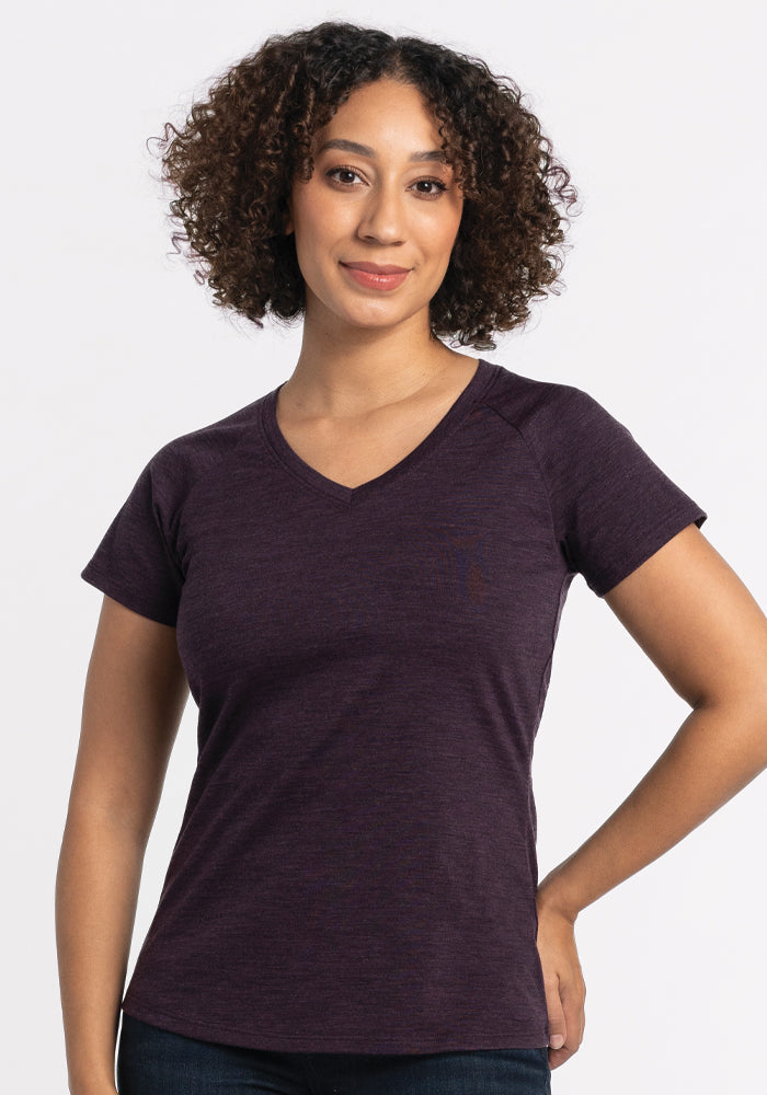 A person with curly hair is wearing a Deep Plum Mia V Neck from Woolx. They are standing against a plain white background, looking forward with a slight smile and their hand on their hip.