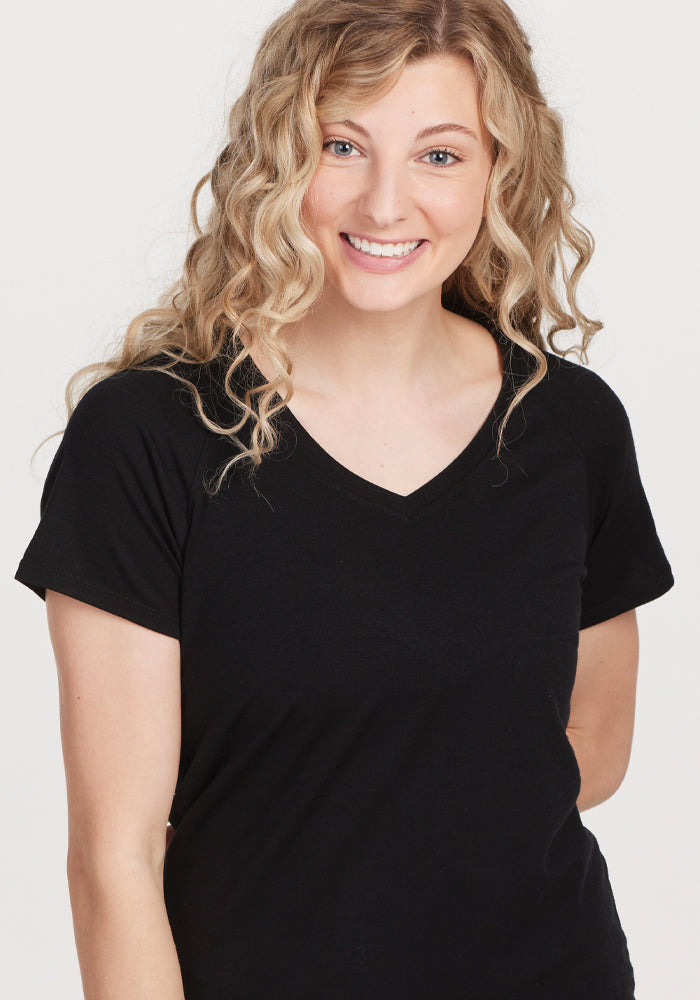 A person with long curly blonde hair is smiling, wearing the Woolx Mia V Neck in black, a high-quality women's T-shirt. They are standing against a plain white background.