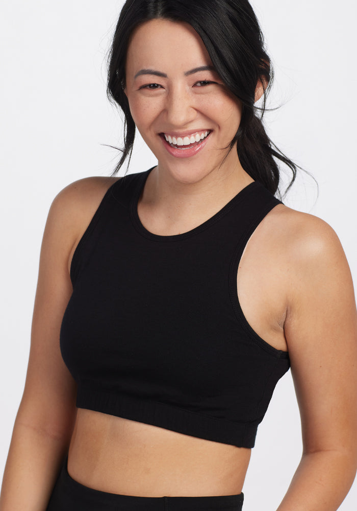 A person with long dark hair smiles in a black sleeveless top, effortlessly paired with the cozy Mila Bra - Black by Woolx, set against a plain background.