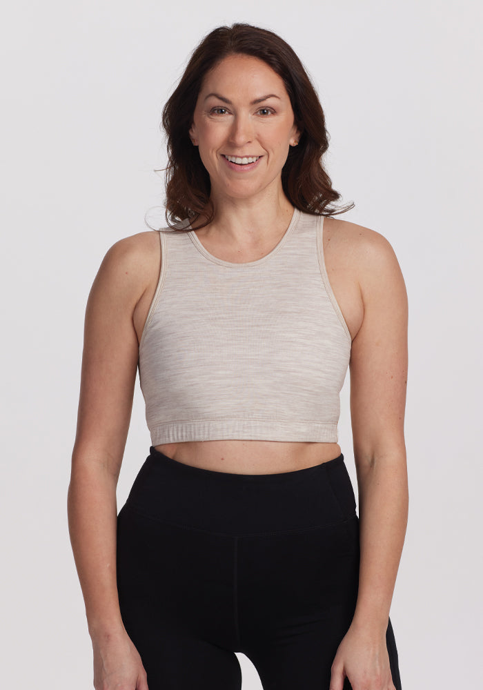 A person with long brown hair, wearing a beige sleeveless top and black leggings, smiles confidently against a white background in a comfortable Woolx Mila Bra - Cream Heather. 