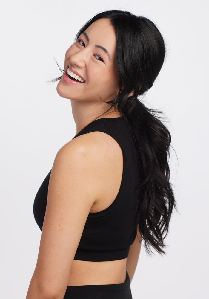 A smiling person with long black hair wears a sleeveless black top, evoking the sleek style of the Woolx Mila Bra - Black. They pose against a white background, playfully glancing over their shoulder at the camera.