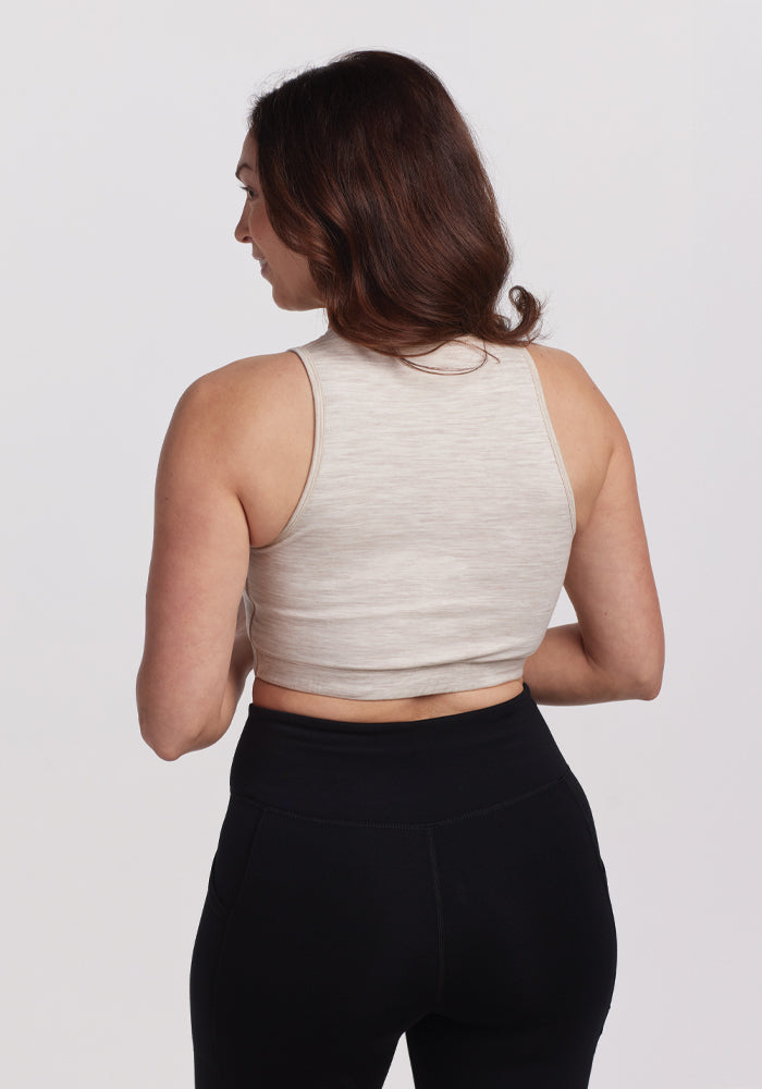 A woman with long brown hair is seen from the back wearing a beige sleeveless top similar to the Woolx Mila Bra in Cream Heather and black leggings against a plain white background.