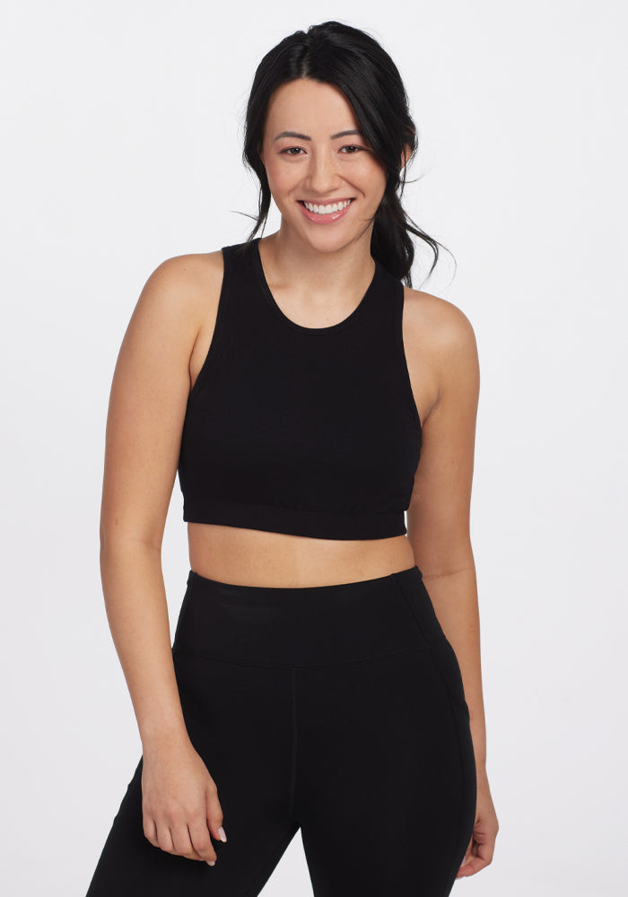 A person with long dark hair smiles, wearing Woolx's Black Mila Bra along with a black sleeveless crop top and matching high-waisted athletic pants, standing against a plain white background. 