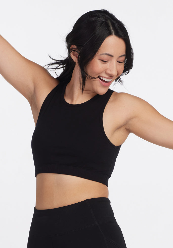 A person with black hair joyfully smiles and raises their arms, wearing the sleek Woolx Mila Bra in black and matching pants. Their confident style against a white background exudes the understated elegance of an Australian Merino Wool blend.