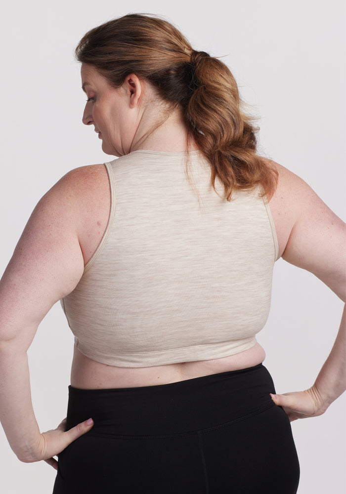 A woman with long brown hair in a ponytail wears the Woolx Mila Bra in Cream Heather and black pants. She faces away from the camera, showcasing her elegant look against a simple white background.