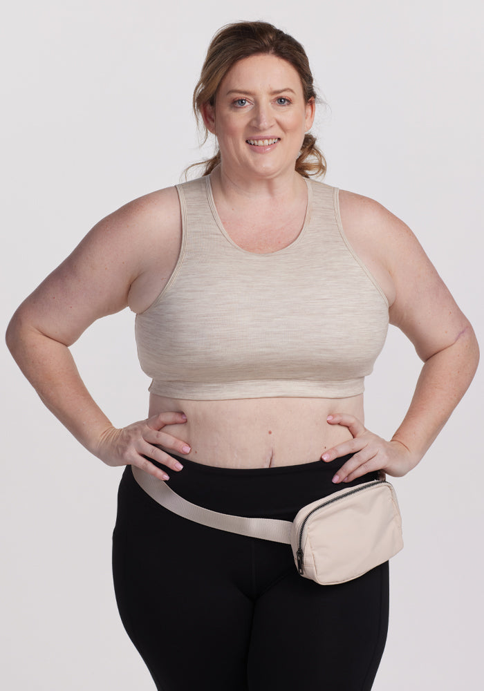 A person wearing a Woolx Mila Bra in Cream Heather smiles at the camera, paired with black leggings and holding a matching beige waist pack, set against a plain, light-colored backdrop. 