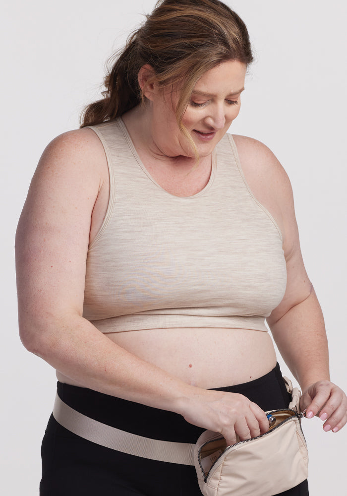 A woman in a beige tank top and black pants smiles while placing a Woolx Mila Bra - Cream Heather into a beige bag, standing against a plain white background.