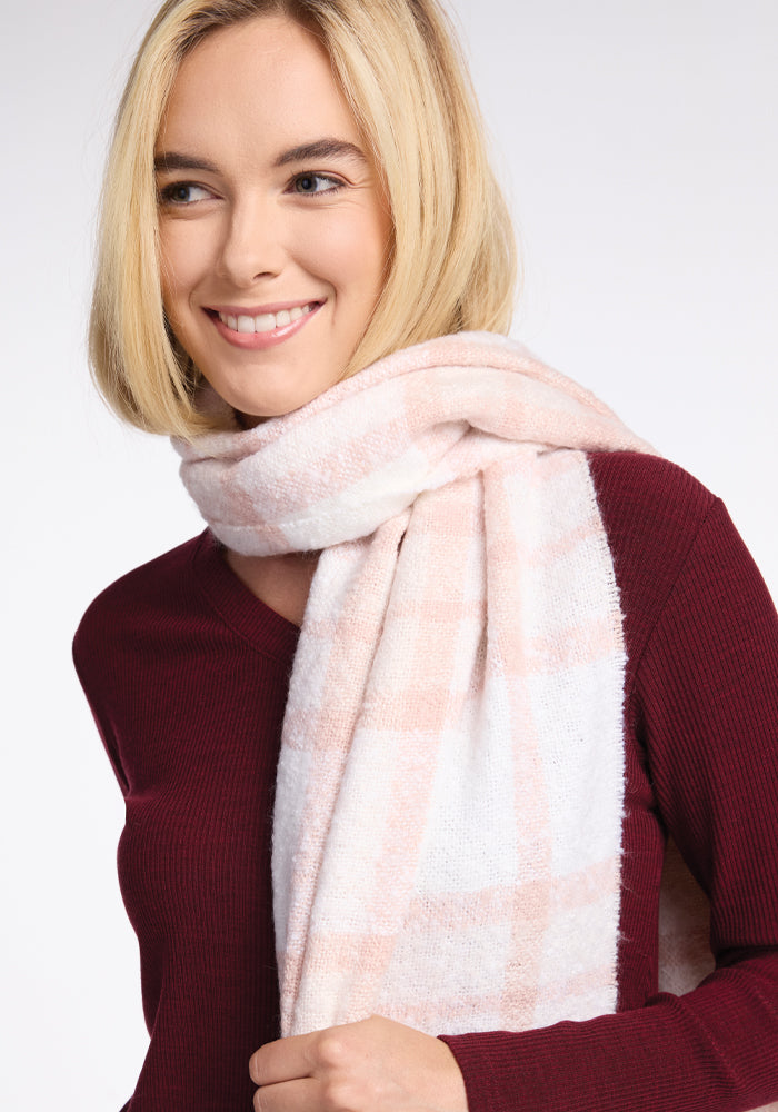 A person with blonde hair smiles while wearing the cozy Bradie Scarf in Mauve Plaid from Woolx and a maroon sweater. The plain white background emphasizes the warmth of the outfit.