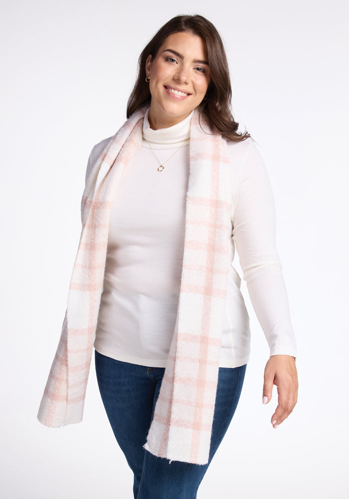 A person wearing a white turtleneck, blue jeans, and the Woolx Bradie Scarf in mauve plaid smiles against a plain white background.