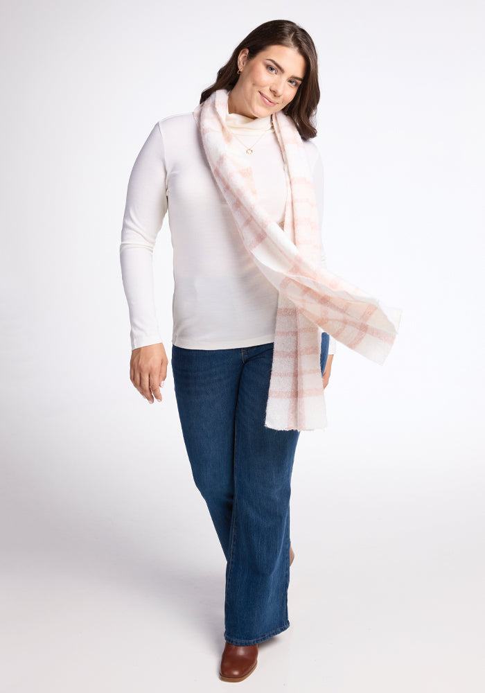 A person is smiling while wearing a white long-sleeve shirt, blue jeans, and brown shoes. They have the Woolx Bradie Scarf in Mauve Plaid draped around their neck and are standing against a plain white background.