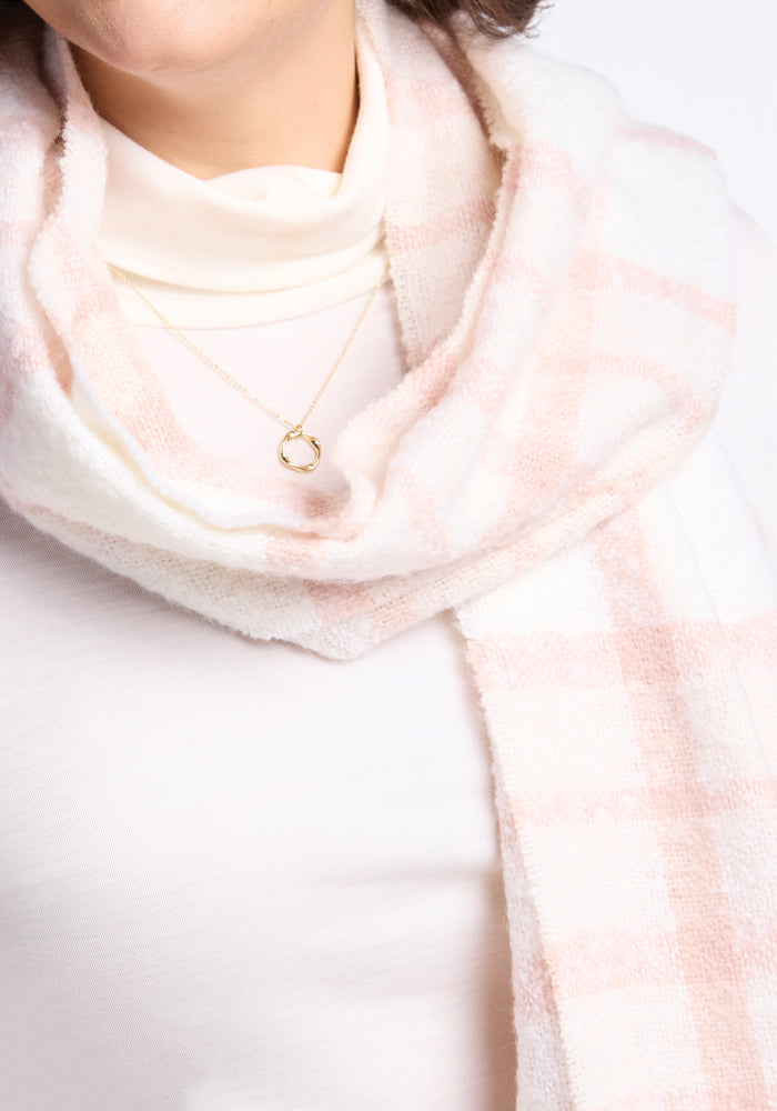 A woman is wearing a soft white turtleneck paired with the Woolx Bradie Scarf in Mauve Plaid, complemented by a delicate gold necklace featuring a circular pendant. The background is white, offering a clean and minimalist appearance.