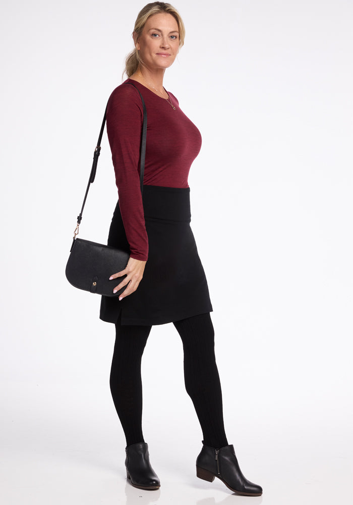 A person stands confidently against a plain backdrop, dressed in a maroon long-sleeve top and the Woolx Monroe Knee Length Skirt in black, crafted from Merino wool. They carry a black shoulder bag and wear ankle boots, with a neutral expression and hair tied back.