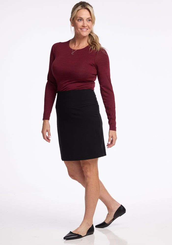 A woman in a maroon long-sleeve top and Monroe Knee Length Skirt in black by Woolx smiles as she poses against a white background. She is wearing black flats, and her blonde hair is styled in a ponytail, complementing her chic merino wool skirt.