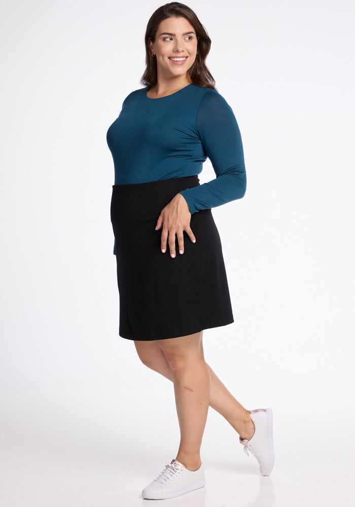 A person smiles while standing with one hand on their hip, exuding knee-length elegance. They are wearing a long-sleeve teal top, the Monroe Knee Length Skirt in black from Woolx made of merino wool, and white sneakers. The background is plain white.