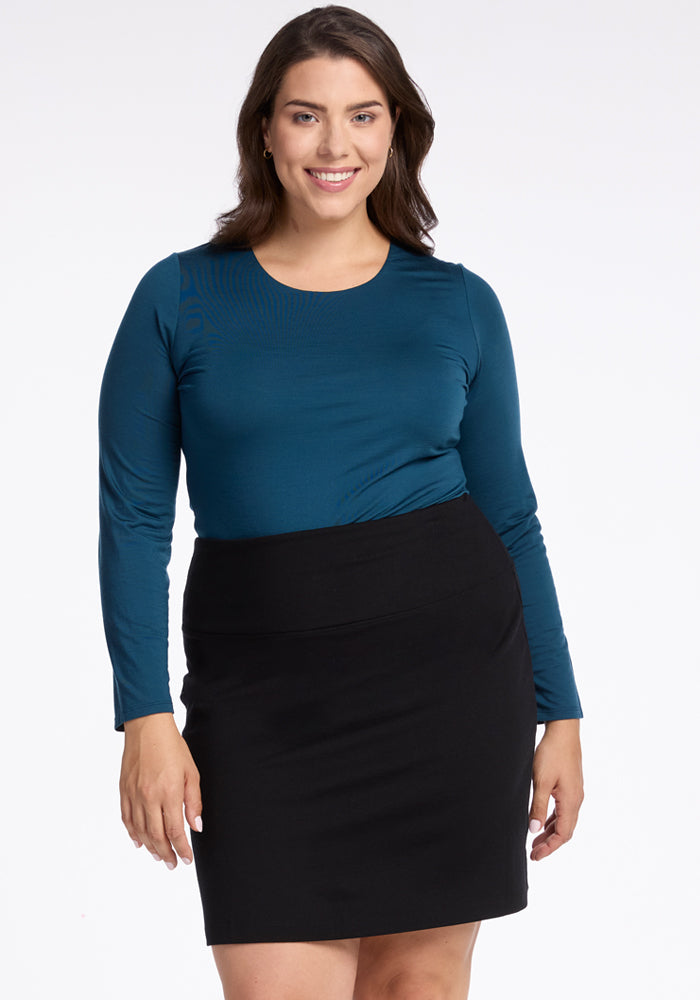 A woman with long brown hair smiles while standing, wearing a teal long-sleeve top and the Monroe Knee Length Skirt in black from Woolx. The background is plain white.