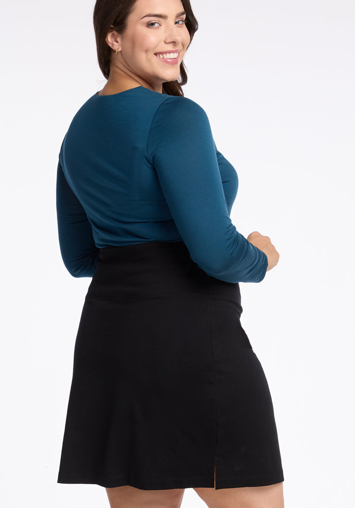 A woman with long dark hair smiles while looking over her shoulder, showcasing the elegance of the Monroe Knee Length Skirt in black from Woolx and a blue long-sleeve top. Her attire is as travel-friendly as it is stylish against the plain white background.