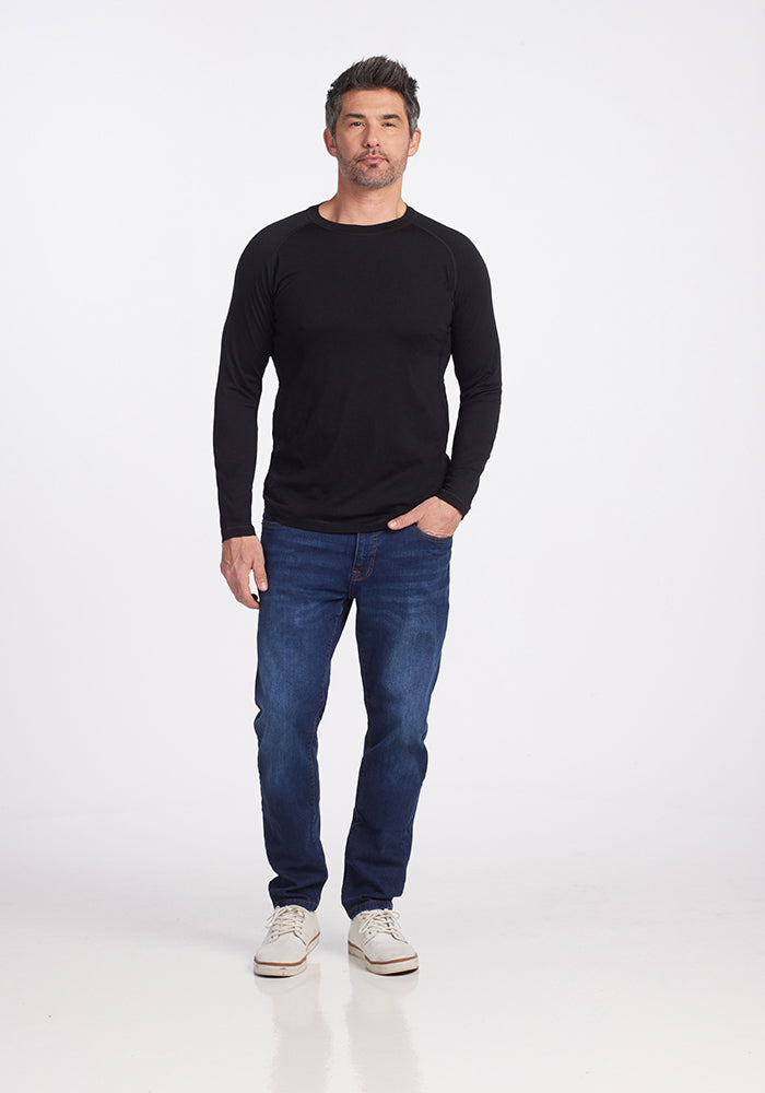 A man stands against a plain white background, wearing the Woolx Essential Tee in black. He pairs it with blue jeans and white sneakers, his right hand resting in his pocket as he gazes forward with a neutral expression.