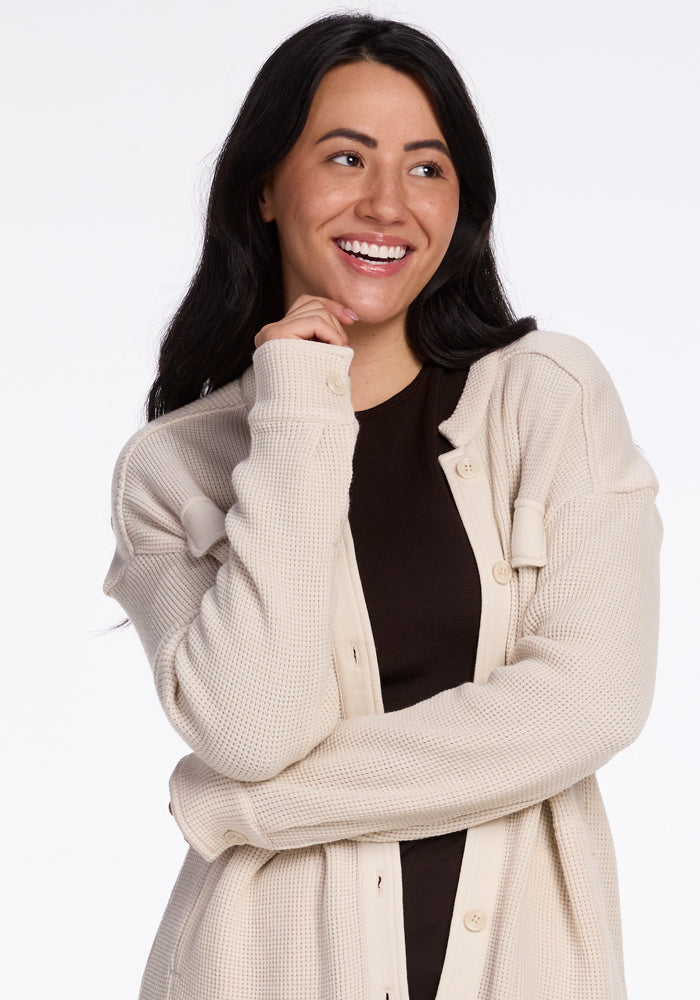 A person with long dark hair smiles while looking off to the side, wearing a Woolx Oaklynn Waffle Shacket in oatmeal over a dark shirt, standing in front of a plain white background.