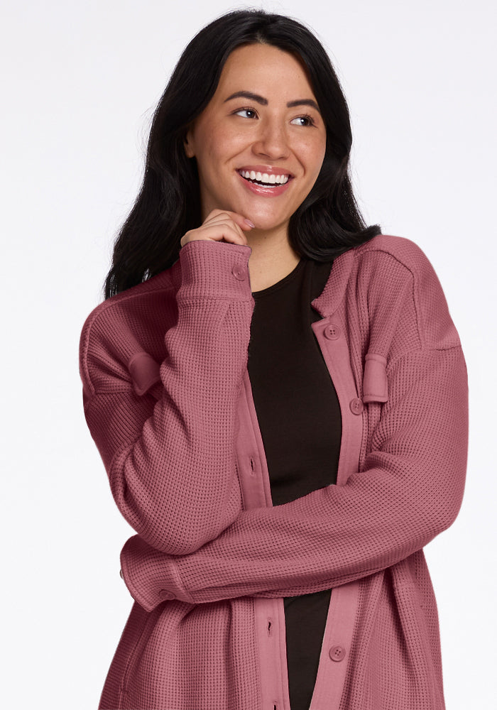 A person with long dark hair, dressed in a Woolx Oaklynn Waffle Shacket in the Wild Ginger shade over a black top, smiles while looking to the side. They are resting their hand on their chin against a plain white background.