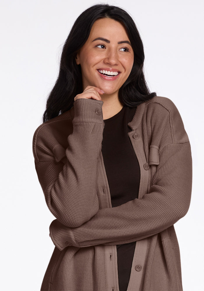 A person with long black hair is wearing the Oaklynn Waffle Shacket in Latte from Woolx over a black shirt. They're smiling and gazing to the side, with one hand thoughtfully touching their chin against a plain white background.